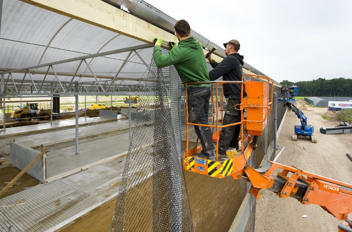 Hier zijn twee bouwers druk in de weer om het vogelgaas in de zijkanten van de serrestal te monteren.